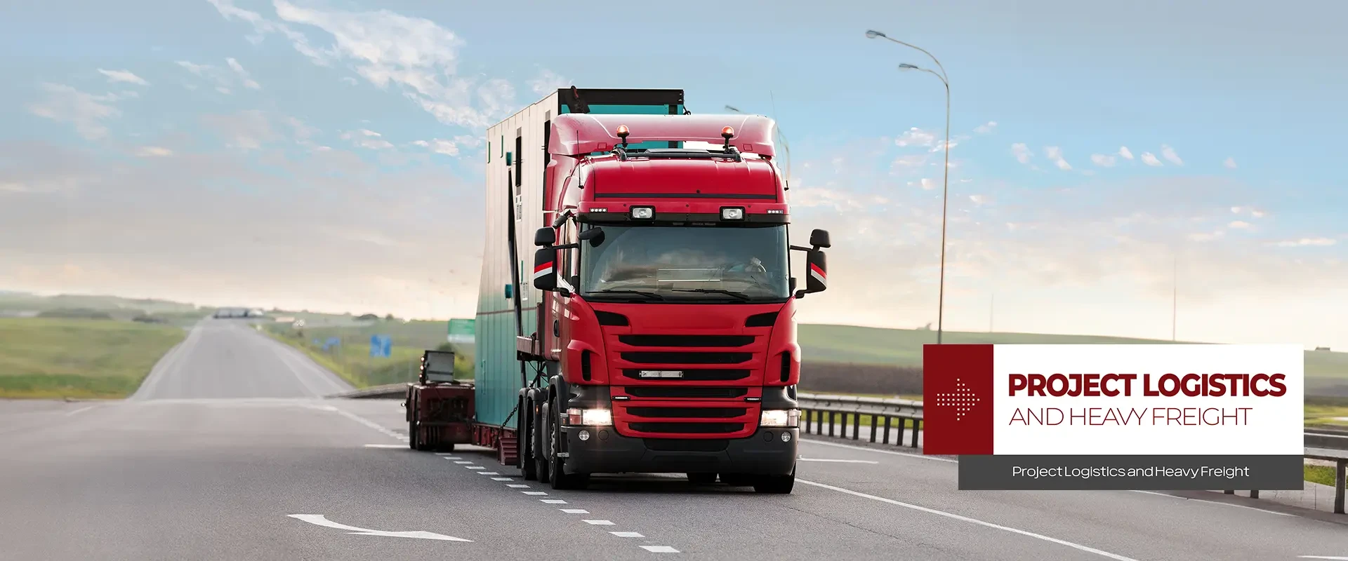 A red truck with a lowbed that says "Project and Heavy Load Transportation" on it is moving on the highway